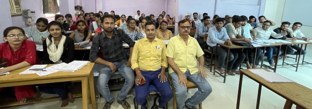 A classroom filled with students seated at desks, with a few students in the foreground interacting with a teacher.